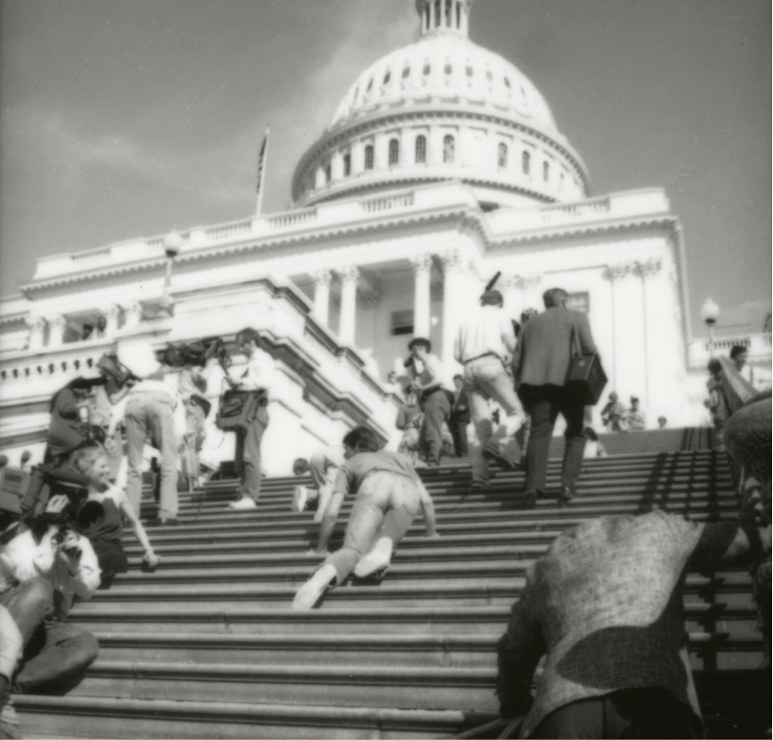 A black and white photo of the protest. Activists are sprawled on the steps of the Capitol Building, some dragging their legs behind them as they climb. Journalists and camera crews are walking around them.