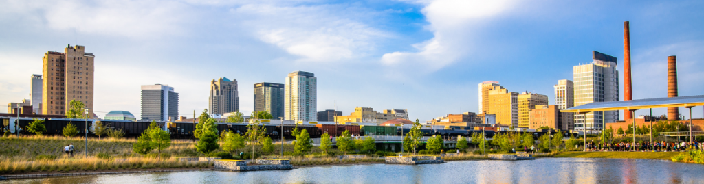 Birmingham city skyline at sunset, with modern high-rises and historic industrial smokestacks reflected in the water at Eastside City Park. (edited) 
