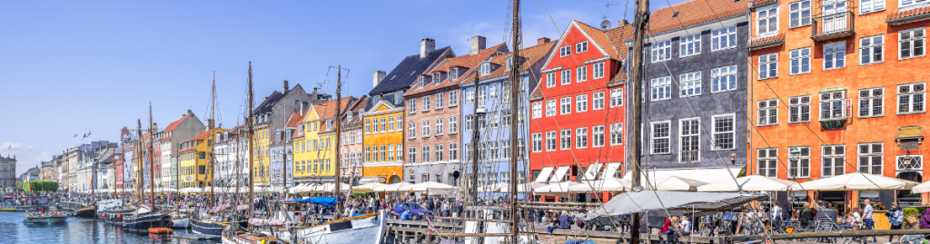 Panoramic view of Copenhagen’s Nyhavn district, featuring rows of brightly painted houses, sailing boats in the canal, and bustling outdoor cafés on a sunny day.
