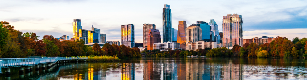 Austin skyline at sunset, with high-rise buildings reflecting golden light over Lady Bird Lake and trees along the shoreline.
