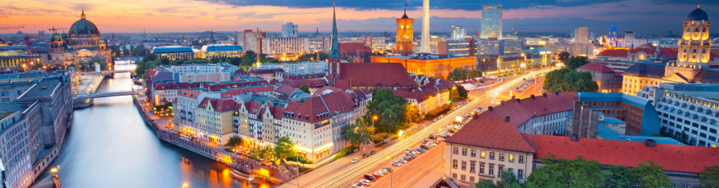 Panoramic view of Berlin at dusk, with city lights reflecting on the Spree River, the Berlin Cathedral dome, and the TV Tower lit against a blue and orange sky.
