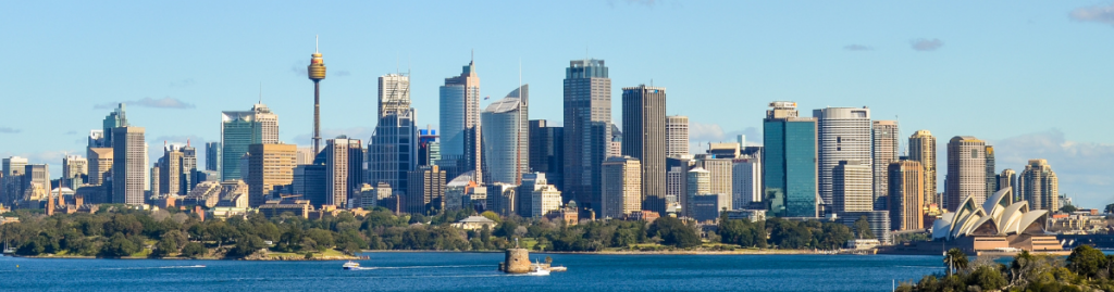 Panoramic view of Sydney, with the Opera House and tall city skyscrapers rising along the waterfront beneath a clear blue sky.
